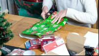 A woman, sitting at a table, wraps a christmas present in brightly coloured christmas wrapping paper. She's trimming the paper t