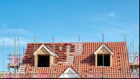 Scaffold poles surrounding the partly tiled roof with dormer windows of a new bungalow being built, against a blue sky.
